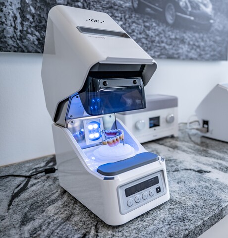 Detailaufnahme des weißen Labolight DUO Polymerisationsgeräts von GC, in dem ein fertiger Zahnersatz mit Zahnfleisch-Basis unter blauem Licht aushärtet. Das Gerät steht auf einer hellgrau gesprenkelten Arbeitsplatte.