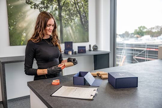 Junge Mitarbeiterin in schwarzer Kleidung und Handschuhen, die einen Oberkiefer-Zahnersatz mit braunem Packpapier umwickelt. Auf dem Tisch liegt ein Lieferschein und eine blaue Verpackungsbox.