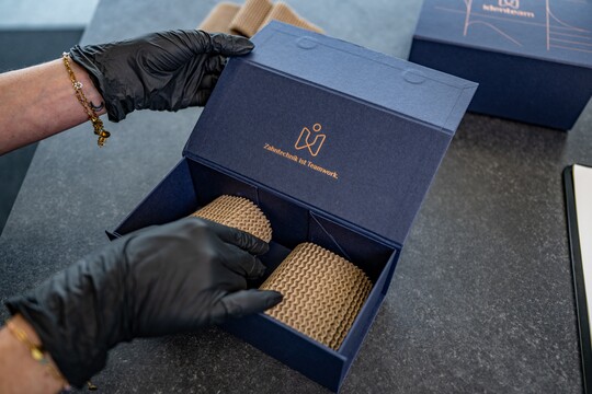 Nahaufnahme der Hände einer Person in schwarzen Schutzhandschuhen, die eine dunkelblaue Transportbox öffnet. Im Deckel steht in kupferfarbener Prägung das Logo und der Spruch "Zahntechnik ist Teamwork."